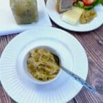 Onion marmalade in a small white bowl on a white plate with a spoon. There is a glass jar of onion marmalade in the background.