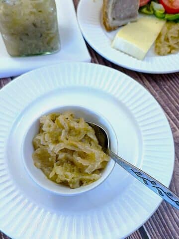 Onion marmalade in a small white bowl on a white plate with a spoon. There is a glass jar of onion marmalade in the background.
