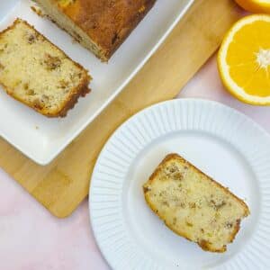 A slice of orange walnut drizzle cake on a small white plate, next to another slice cut from the cake on a white platter.