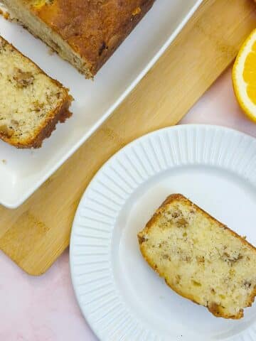 A slice of orange walnut drizzle cake on a small white plate, next to another slice cut from the cake on a white platter.