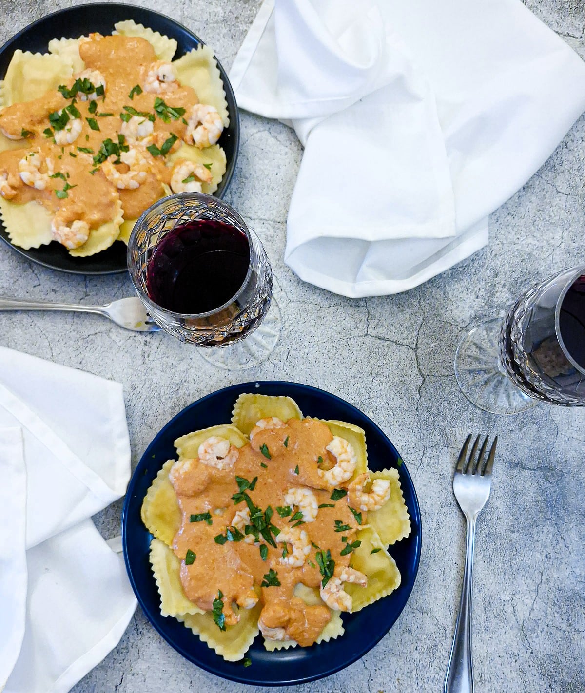Two plates of ravioli and red pepper sauce with white serviettes and glasses of red wine.