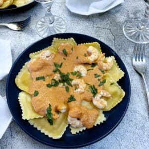 A blue dinner plate with pieces of ravioli topped with red pepper sauce and garnished with prawns.