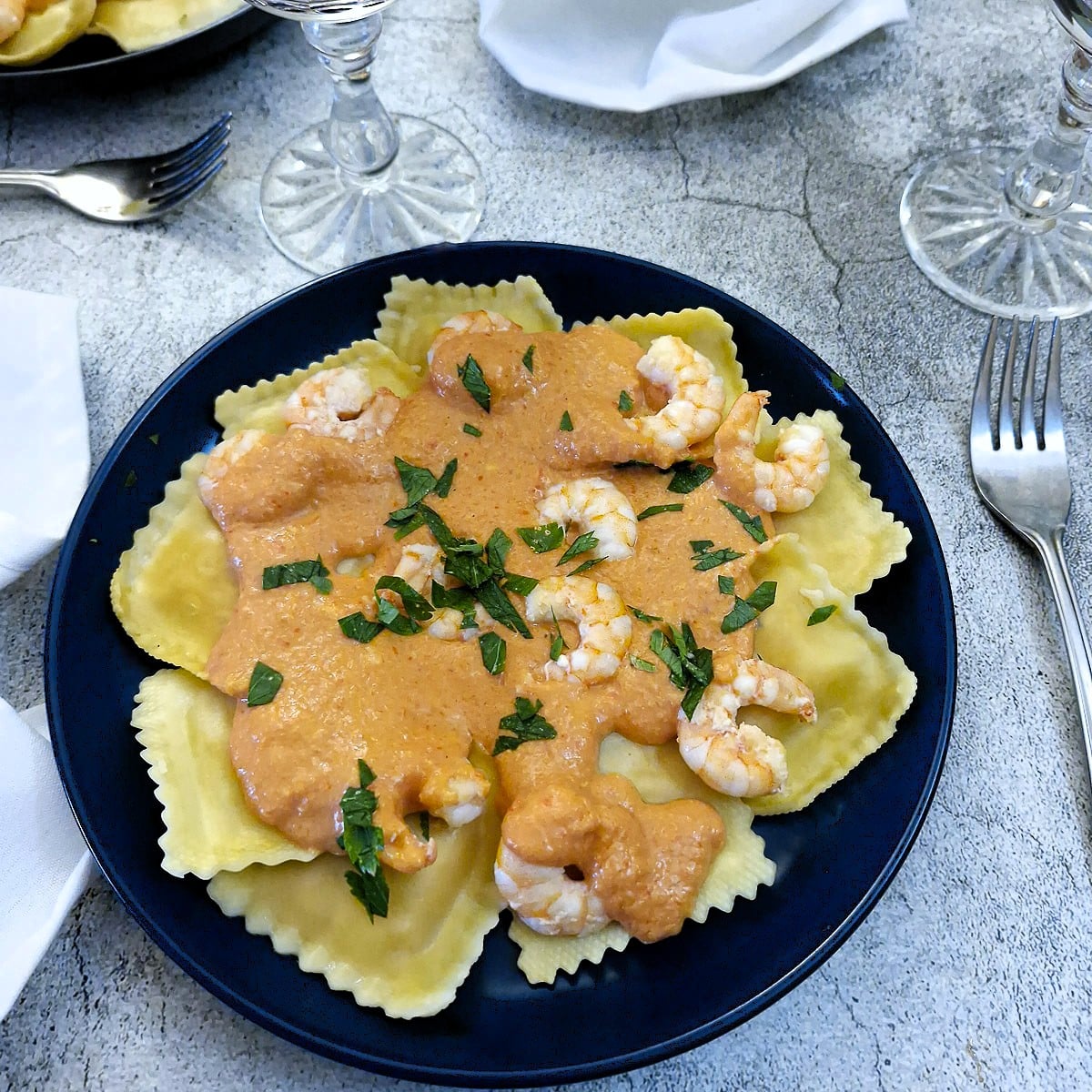 A blue dinner plate with pieces of ravioli topped with red pepper sauce and garnished with prawns.