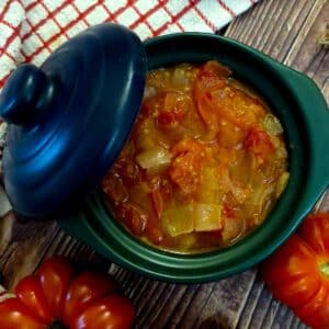 A green serving dish filled with delicious tomato and onion stew. The lid of the dish is propped on the side.