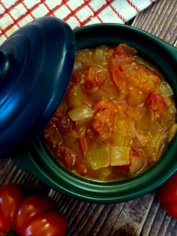 A green serving dish filled with delicious tomato and onion stew. The lid of the dish is propped on the side.