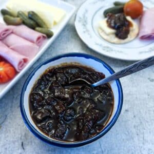 A small dish of marrow and apple chutney with a teaspoon for serving. There is a platter of cold meat and cheese in the background.