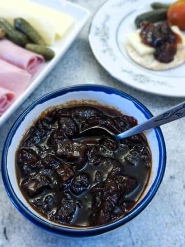 A small dish of marrow and apple chutney with a teaspoon for serving. There is a platter of cold meat and cheese in the background.