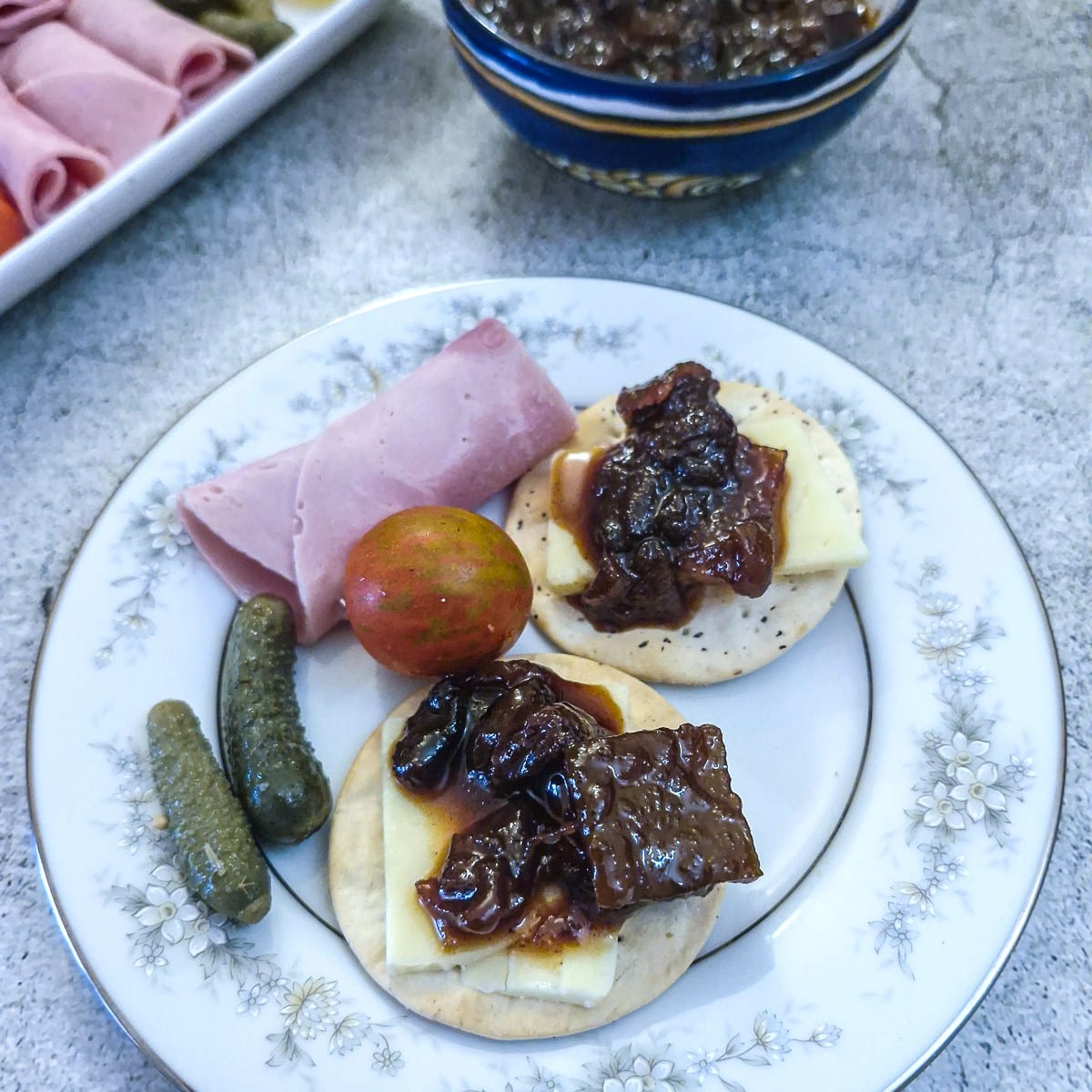 Two crackers topped with cheese, with a spoonful of marrow an apple chutney on top. There is also a slice of ham, gherkins and tomato on the plate.