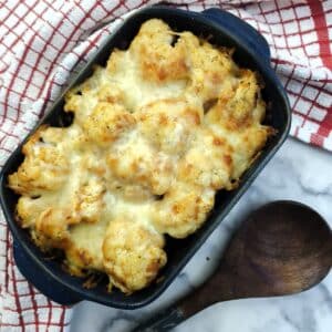 A blue dish of cheesy cauliflower tomato bake on a table with a large wooden spoon. There is a red and white checked cloth around the dish.