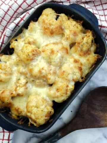 A blue dish of cheesy cauliflower tomato bake on a table with a large wooden spoon. There is a red and white checked cloth around the dish.