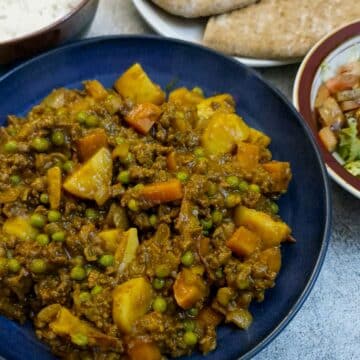 A blue serving dish of beef mince curry in a hurry. There is a bowl of rice and a few slices of pita bread in the background.