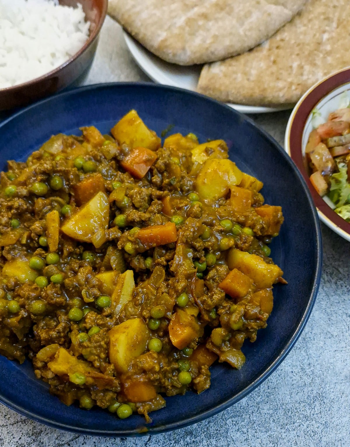 A blue serving dish of beef mince curry in a hurry. There is a bowl of rice and a few slices of pita bread in the background.