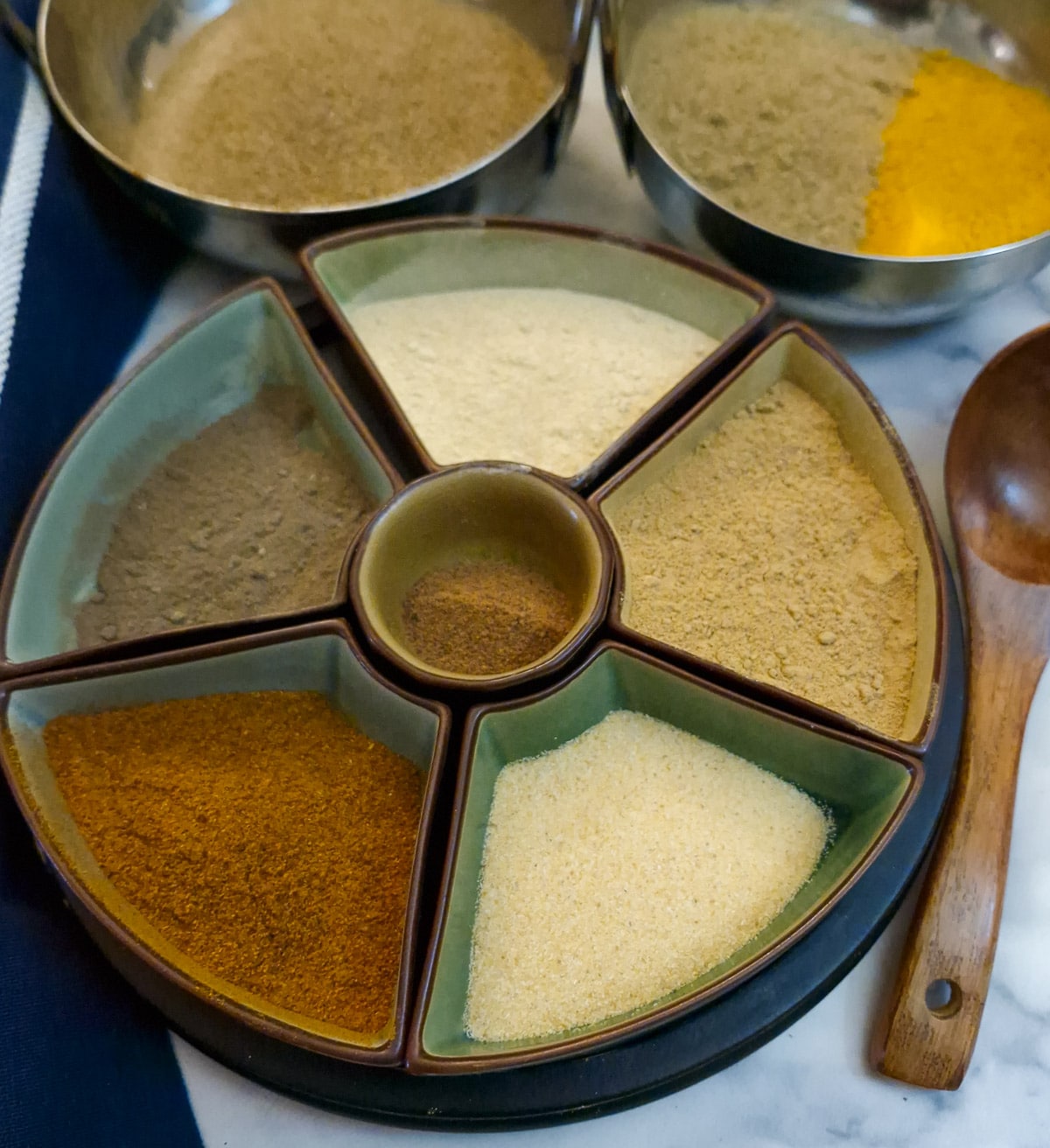 Various bowls on spices on a table with a wooden spoon.