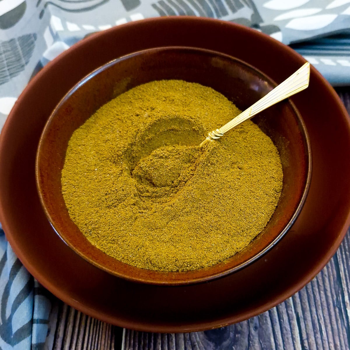 A brown dish of homemade curry powder. There is a small gold-plated spoon in the dish.