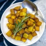 A white pllate of cubed roasted butternut topped with a sprig of rosemary.