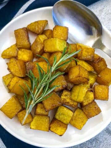 A white pllate of cubed roasted butternut topped with a sprig of rosemary.