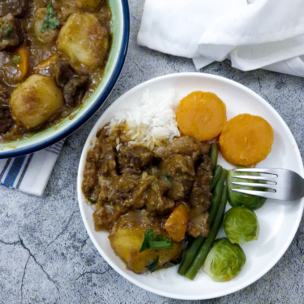 Tamatie bredie spooned over a serving of white rice on a white plate with slices of butternut and a few green beans.