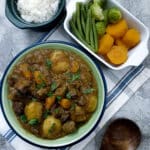 A green serving dish of tamatie bredie, with dishes of vegetables and rice on the table.