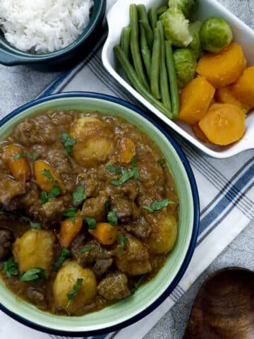 A green serving dish of tamatie bredie, with dishes of vegetables and rice on the table.
