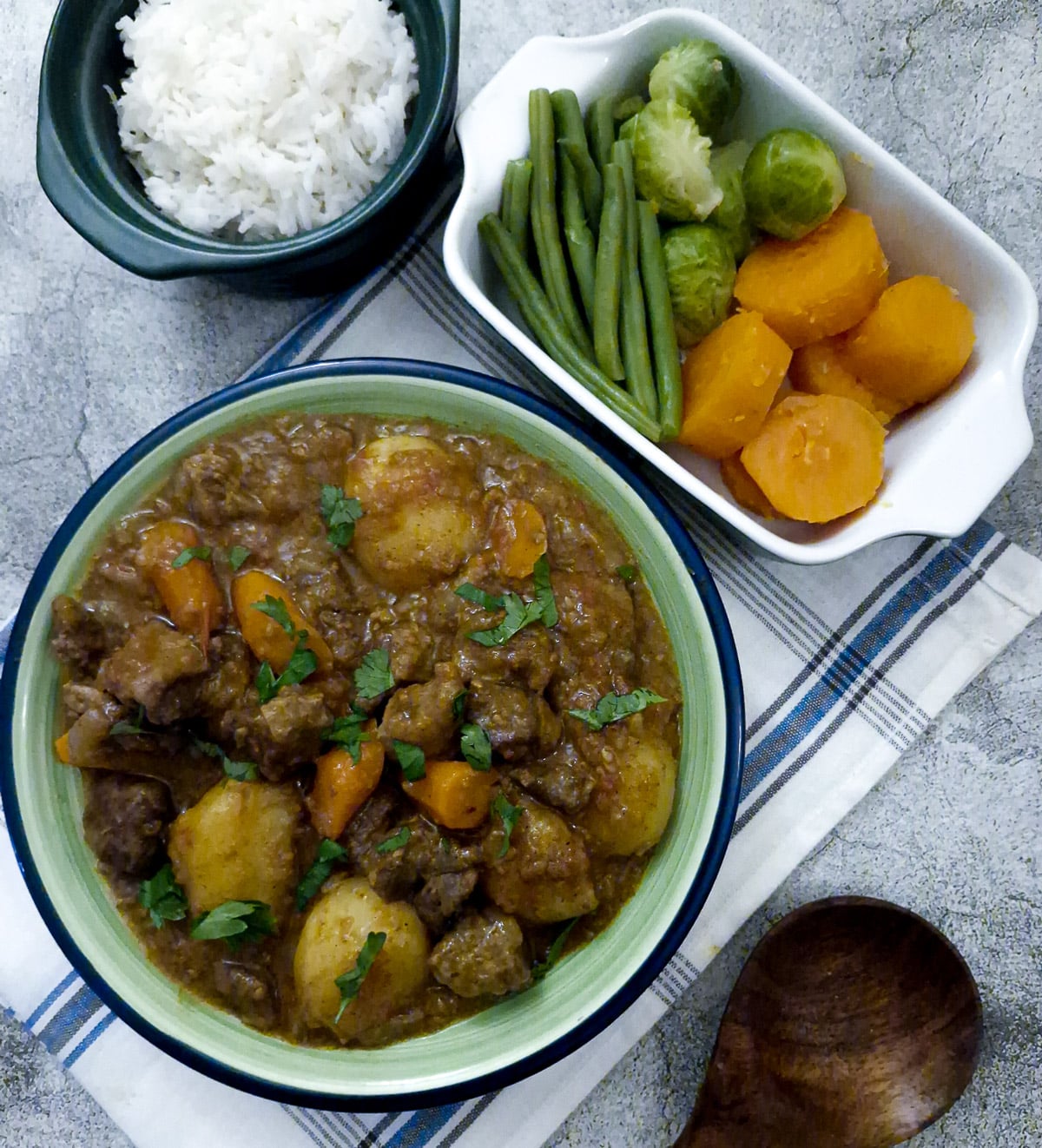 A green serving dish of tamatie bredie, with dishes of vegetables and rice on the table.