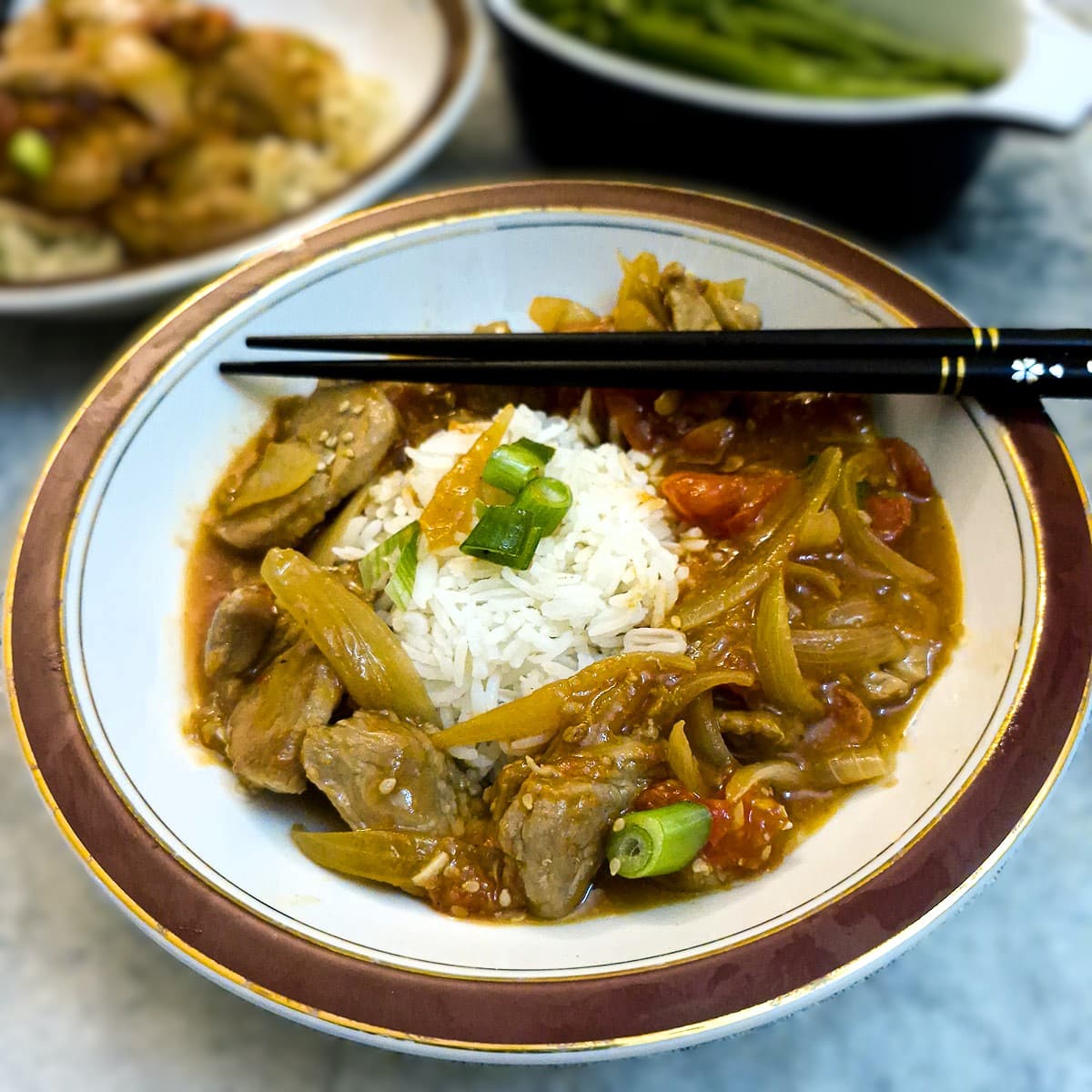 Pork and tomato stir-fry in a red-rimmed dish, spooned alongside a mound of white rice. There is a pair of black chopsticks resting on the bowl.