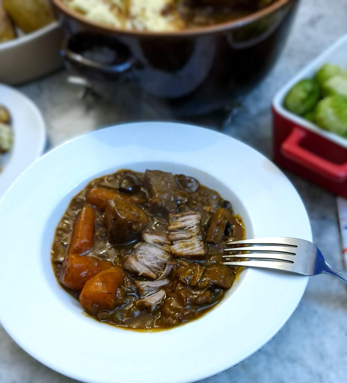 A white dinner plate with a helpingt of French onion beef stew. There are dishes of vegetables on the table.