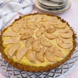 A round baked pear and ginger tart on a glass plate. There are slices of pears arranged on top of the custard filling.