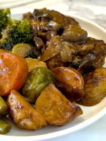 A dinner plate with 2 patties of Salisbury steak smothered with mushroom and onion gravy, with a selection of potatoes and vegtables on the side.