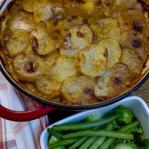 A dish of steak and kidney hotpot, topped with golden, crispy sliced potatoes. There is a dish of green beans in the foreground.