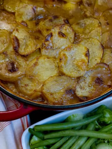 A dish of steak and kidney hotpot, topped with golden, crispy sliced potatoes. There is a dish of green beans in the foreground.