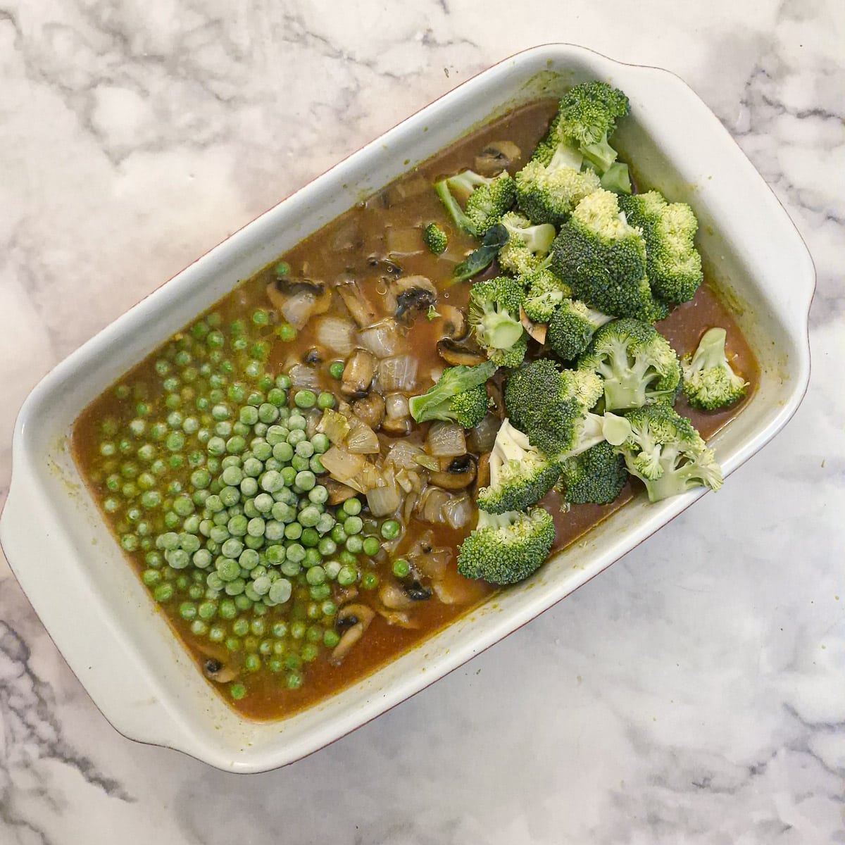 Peas and broccoli florets added to the rice and stock in the white ceramic baking dish.