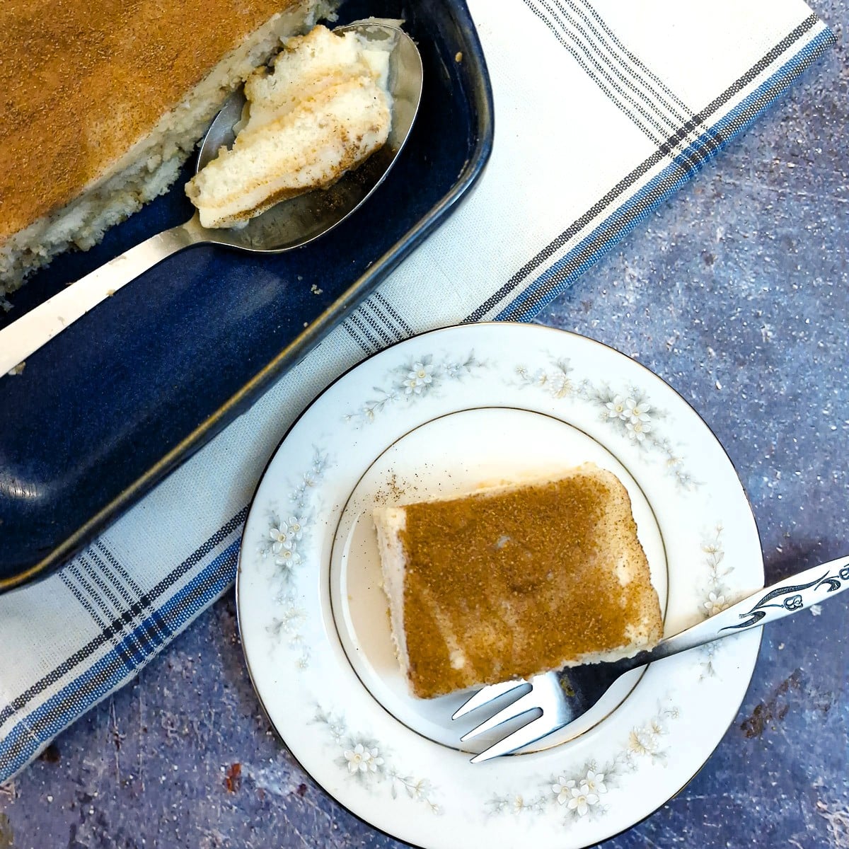 A slice of Baileys milk tart tiramisu on a small dessert plate. There is a dish of tiramisu in the top corner of the photo with a serving spoon filled with tiramisu.