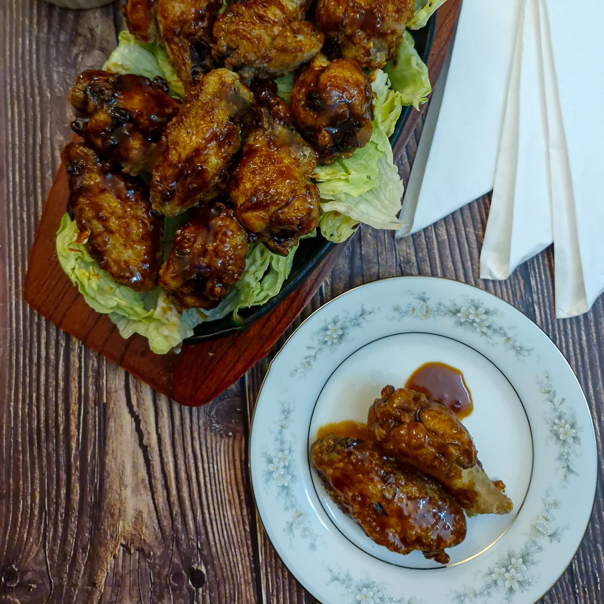 Two sauce-covered chicken wings on a small plate, next to a platter of chicken wings.