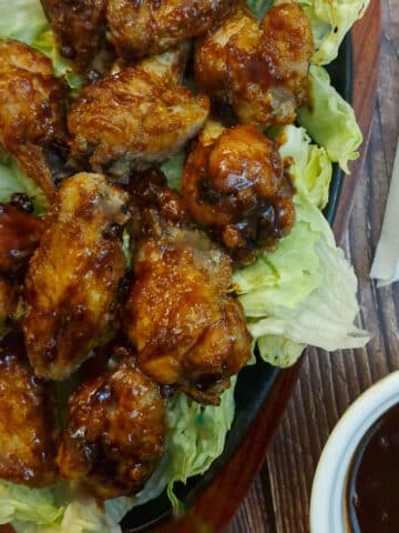 A pile of gochujang chicken wings on bed of lettuce leaves. There is a small dish of extra sauce in the foreground.