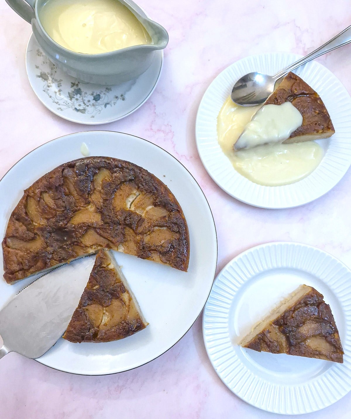 A caramelised frying pan apple cake on a serving plate. There is a green jug of custard in alongside, with 2 side plate holding slices of the cake.