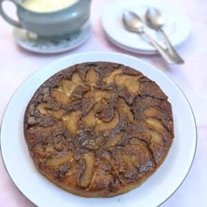 A fryuing pan apple cake tipped out of the pan onto a white plate. The caramelised apples and brown sugar that were on the bottom of the pan now form the top of the cake.