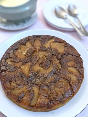 A fryuing pan apple cake tipped out of the pan onto a white plate. The caramelised apples and brown sugar that were on the bottom of the pan now form the top of the cake.