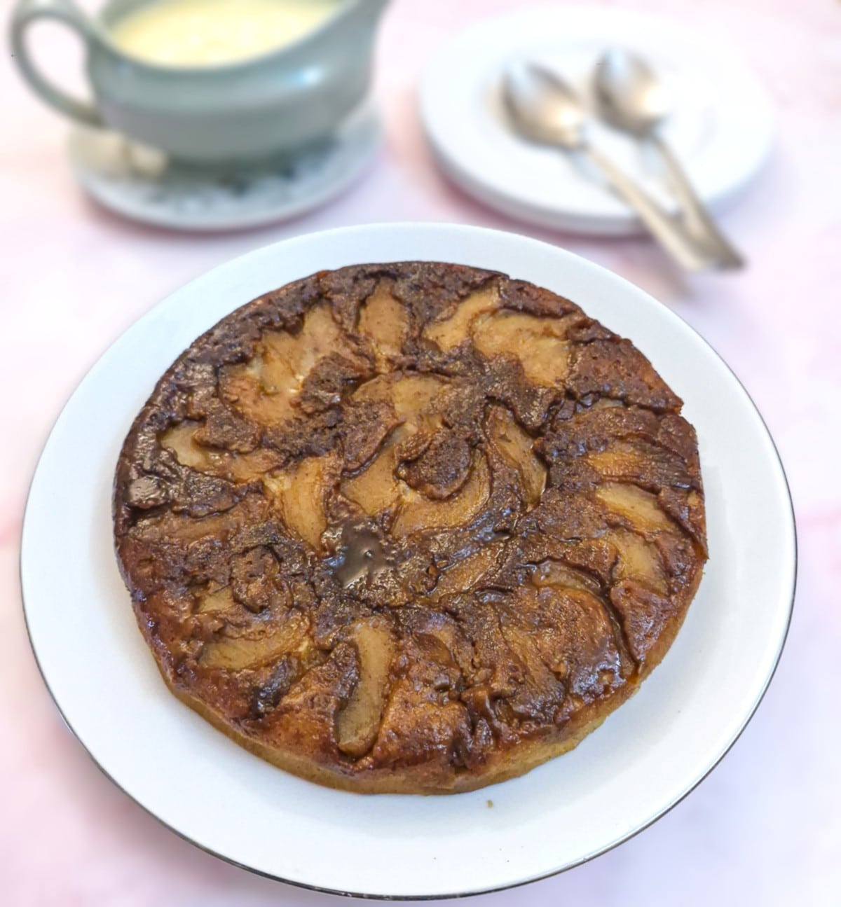 A fryuing pan apple cake tipped out of the pan onto a white plate.  The caramelised apples and brown sugar that were on the bottom of the  pan now form the top of the cake.