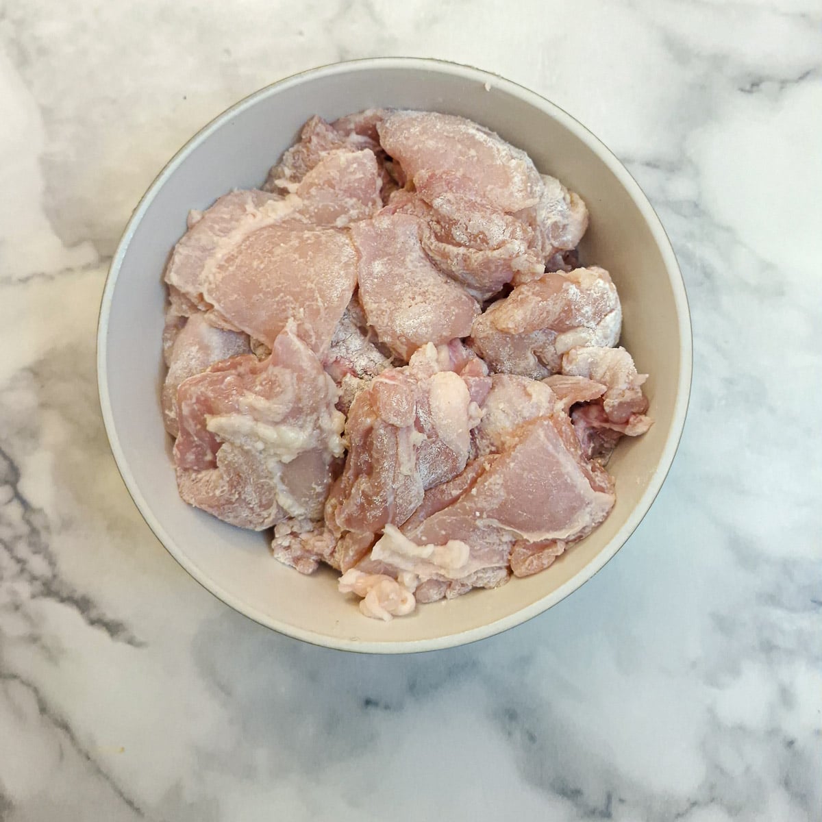 Pieces of chicken thigh fillets lightly coated with flour, in a small white bowl.