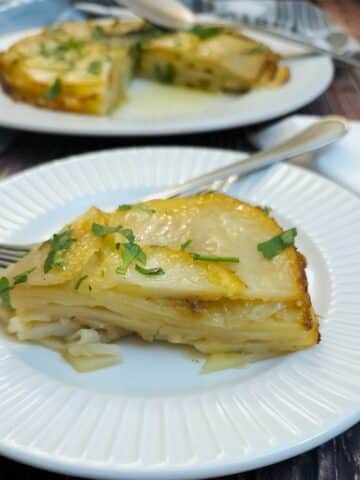 A slice of welsh onion potato cakeon a white plate with a fork. There is the entire cake in the background.