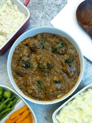 Beef olives in a white serving dish, covered with gravy, garnished with pieces of wilted basil. The serving dish is surrounded by dishes of vegetables and mashed potatoes and there is a large wooden serving spoon alongside.