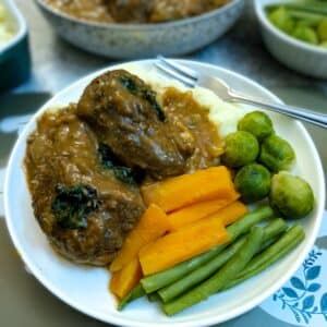 Two beef olives arranged on a bed of mashed potatoes on a white plate with carrots, green beans and brussels sprouts. There is a hearty helping of gravy poured over the beel olives.