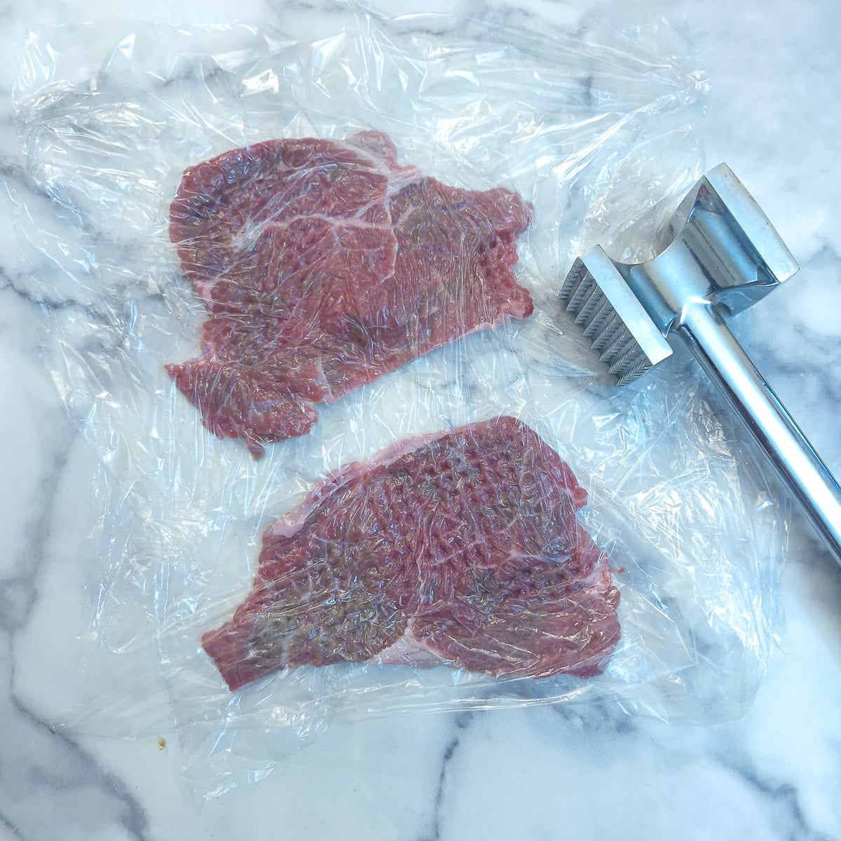 Step 4 - 2 slices of beef enclosed in plastic wrap and being flattened with a meat mallet.