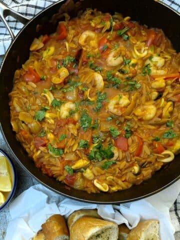 Overhead shot of fideua seafood pasts in a large black frying pan. There are dishes of lemon wedes and garlic bread alongside.