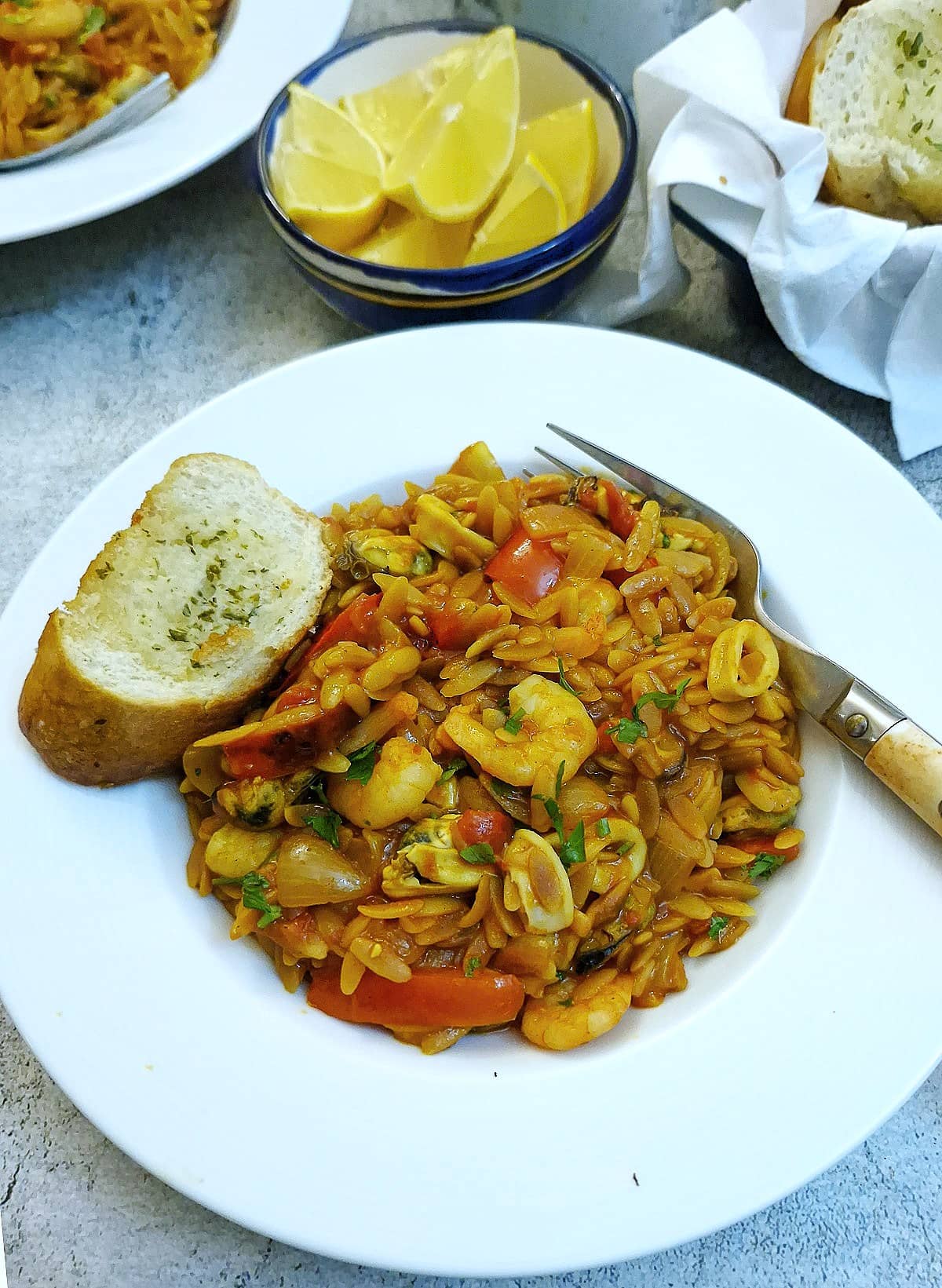 A helping of seafood pasta in a white dish with a slice of garlic bread on the side.  There is a bowl of lemon wedges in the backgroun.