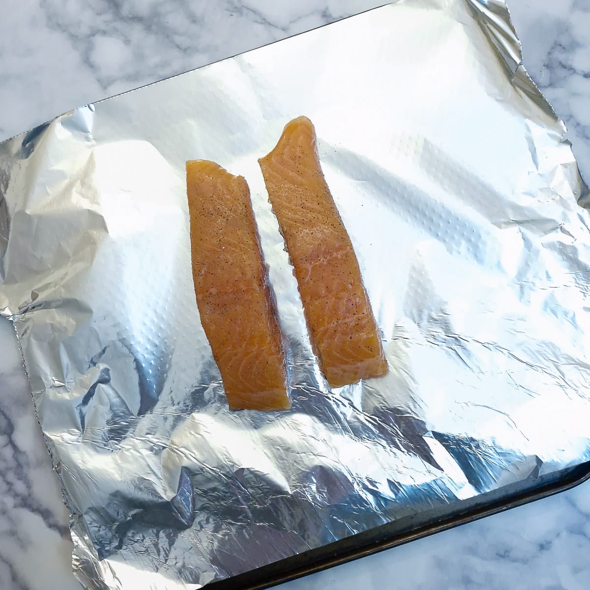 Two pieces of raw salmon on a sheet of tin foil on a baking tray.