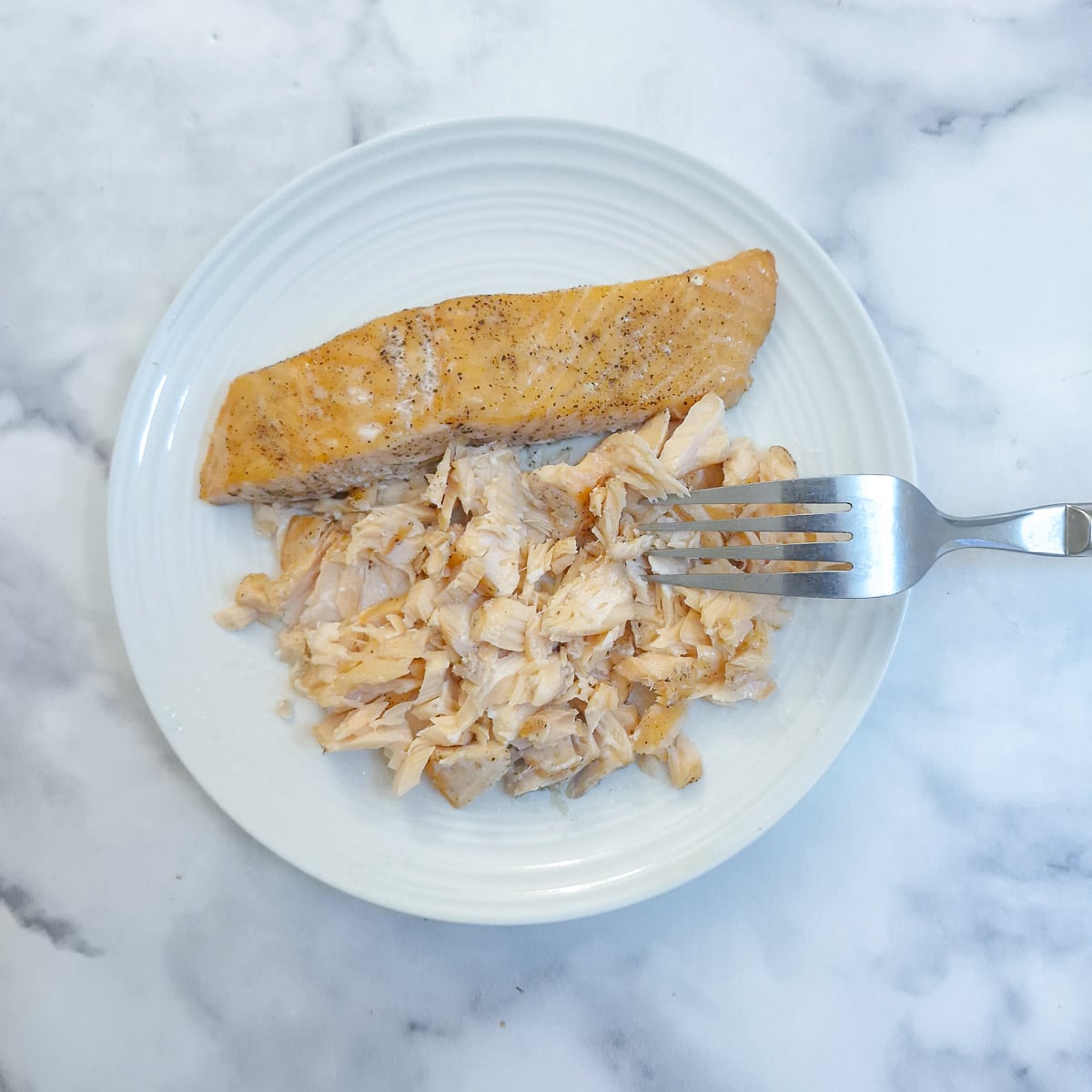 Cooked salmon being flaked with a fork on a white plate.