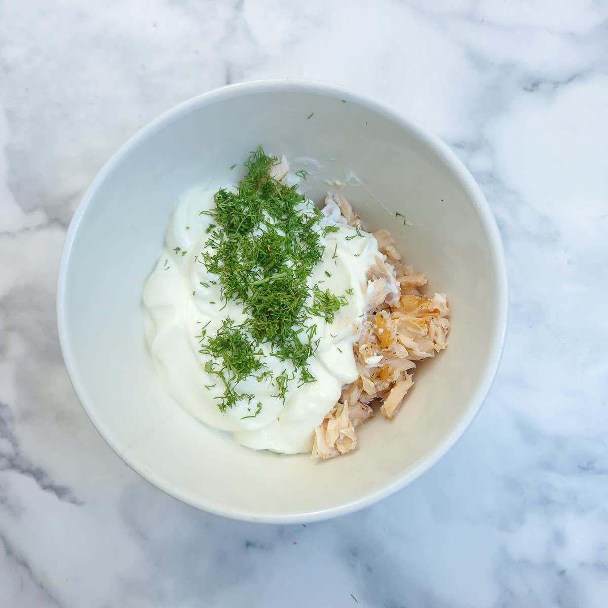 Flaked salmon in a white mixing bowl with mayonnaise, creme fraiche and chopped dill.