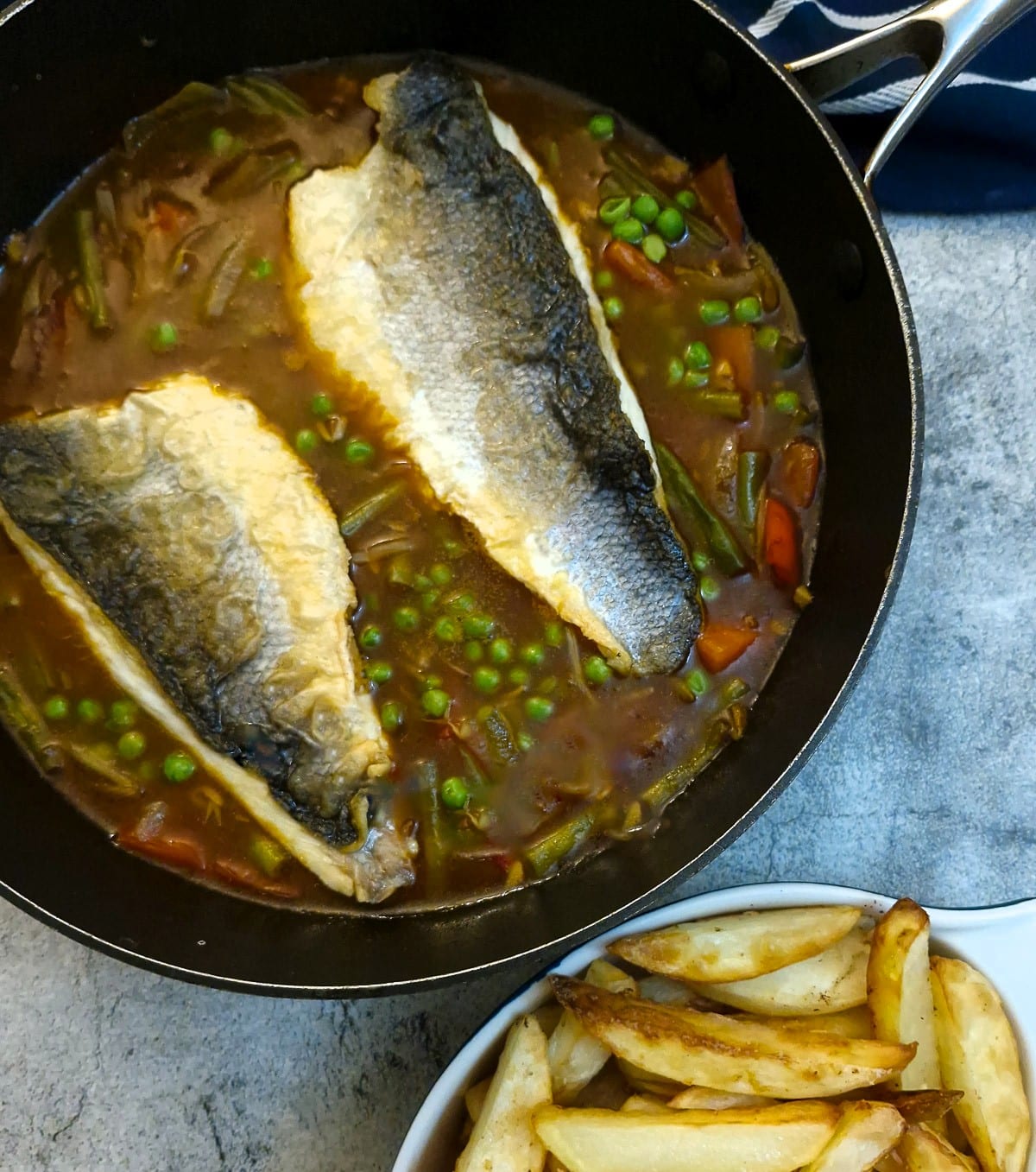 2 fillets of sea bass on top of the tasty garlic qand vegetable sauce in a fryng pan. There is a dish of chips in the foreground.