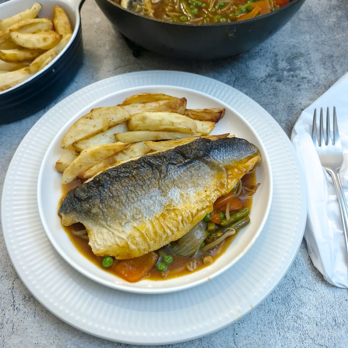 A piece of crispy-skinned sea bass on a dish of garlic vegetable sauce with french fries on the side.